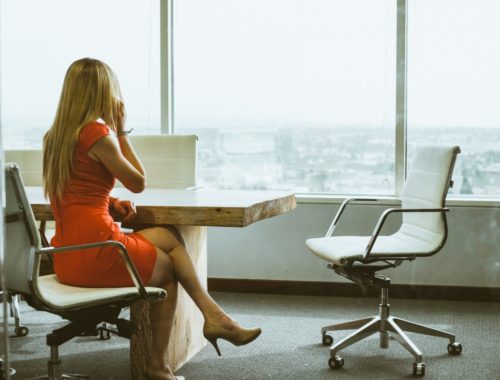 Žena u narančastoj haljini sjedi u uredu s pogledom na grad